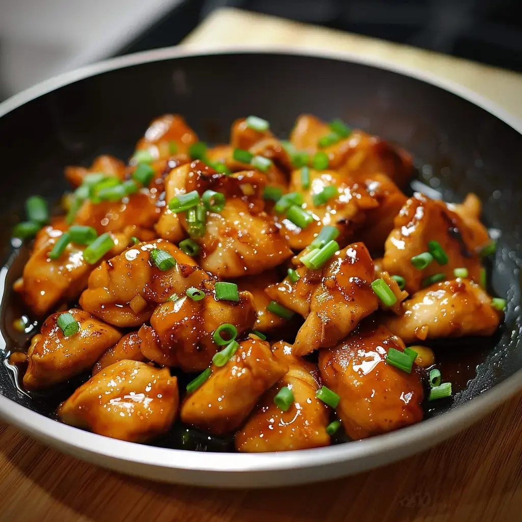 Eine Schale mit gebratenem Huhn in süßer Sauce, getoppt mit gehacktem Schnittlauch, serviert in einer schwarzen Schüssel.
