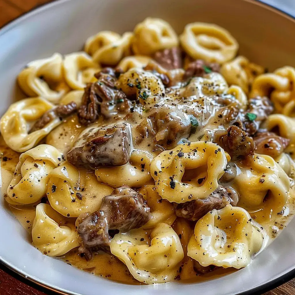 Eine Schüssel mit cremigen Tortellini, Fleischstücken und Gewürzen, appetitlich angerichtet.