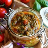 Ein Glasgefäß mit einer mediterranen Pesto-Kreation aus Tomaten, Kräutern und Gewürzen, umgeben von frischen Tomaten und Knoblauchzehen.